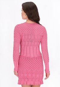 Femme aux longs cheveux bruns portant une robe en maille rose à manches longues avec un motif en losanges, vue de dos sur fond blanc.