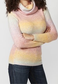 Person with curly hair, arms crossed, wearing a pastel multicolor turtleneck sweater and dark blue jeans against plain background.