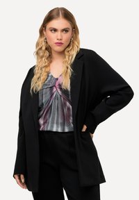 Blazer negro de gran tamaño con mangas largas, usado sobre una blusa plisada multicolor con tonos morados, grises y rosas. Pantalones de corte ajustado.