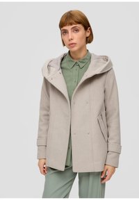Jeune femme aux cheveux courts portant un manteau à capuche beige clair sur une chemise verte à boutons et un pantalon assorti, sur fond uni.