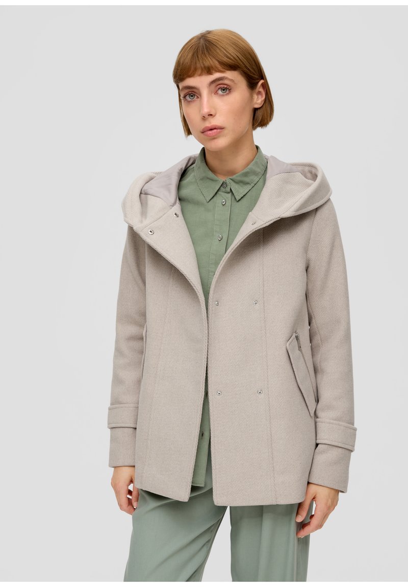 Jeune femme aux cheveux courts portant un manteau à capuche beige clair sur une chemise verte à boutons et un pantalon assorti, sur fond uni.