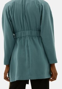 Jasnoniebieski, długi płaszcz z paskiem o luźnym kroju, z długimi rękawami i gładką fakturą. Posiada detal w postaci marszczenia z tyłu.