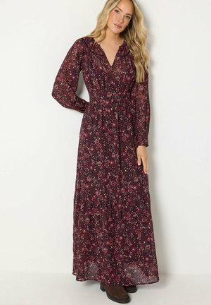 Femme aux longs cheveux blonds porte une robe noire à manches longues avec un imprimé floral rouge et des bottes noires, debout sur un fond clair.