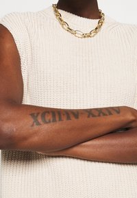 Pletený béžový tílko bez rukávů s vysokým límcem a texturovaným povrchem. Nyní je nošen zlatý řetízek s oválnými články a průhlednými akcenty.