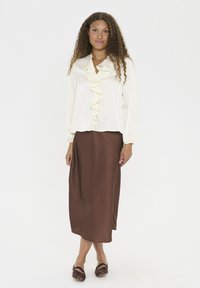 Lichtcreme blouse met een gerimpeld voorpand en lange mouwen, gecombineerd met een lange bruine rok. Bruine schoenen maken de outfit compleet.