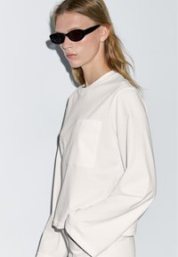 White long-sleeve top with a round neckline and front pocket. Black cat-eye sunglasses. Model poses against a plain background.
