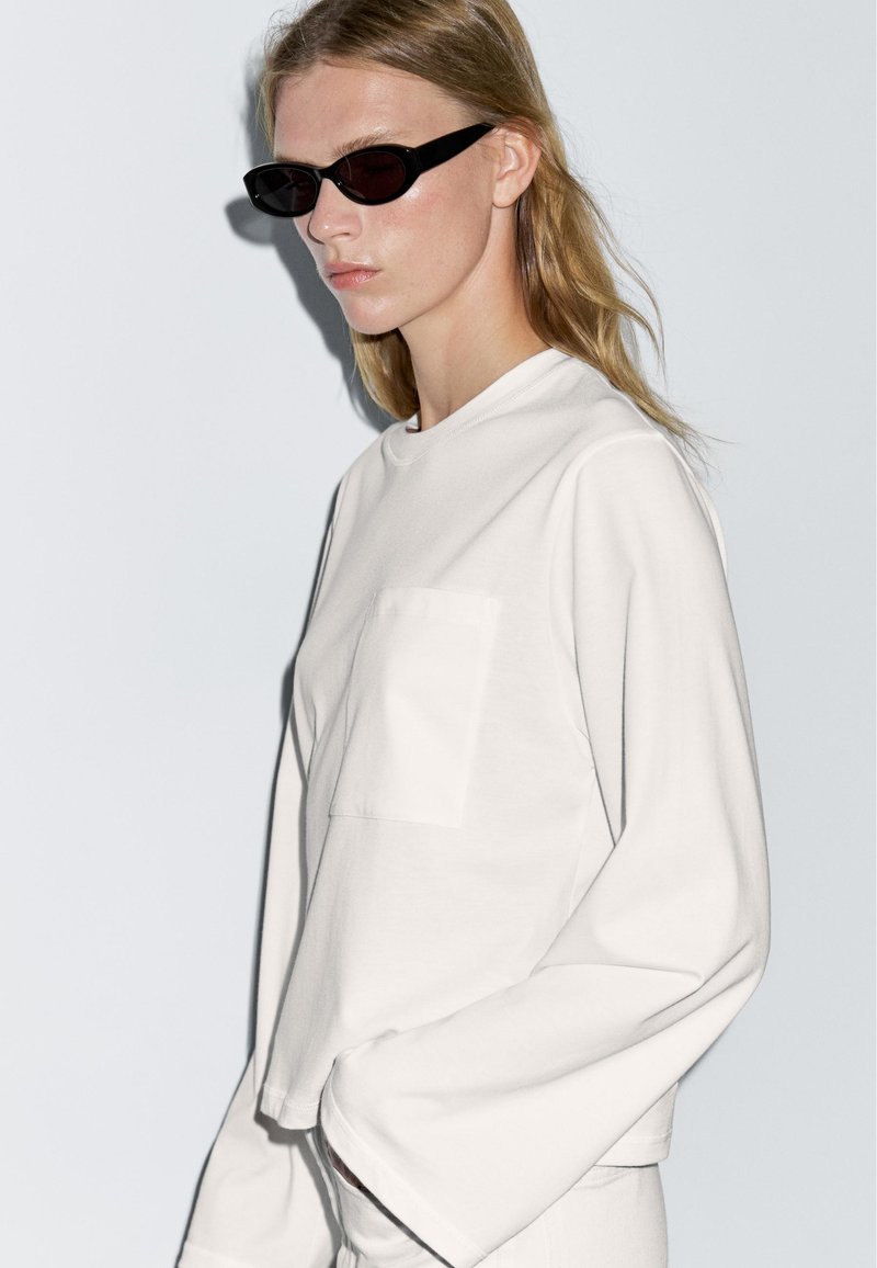 White long-sleeve top with a round neckline and front pocket. Black cat-eye sunglasses. Model poses against a plain background.