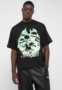 Tricou negru cu mânecă scurtă, prezentând un grafic cu flacără verde și multiple craniene; include textul „+TEAMGEIST” în partea de jos și logo-ul adidas.