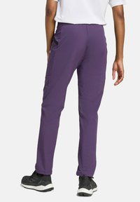 Pantaloni outdoor viola con una texture liscia, dotati di tasche laterali e dettagli neri, abbinati a scarpe da ginnastica nere.