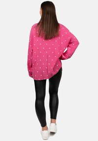 Blusa rosa a trama con motivi a forma di cuore bianchi, abbinata a leggings neri lucidi e sneakers bianche. Visibile vestibilità ampia e maniche lunghe.