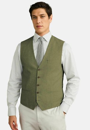 Man wearing green buttoned vest, striped white shirt, patterned gray tie, and light-colored pants against plain background.