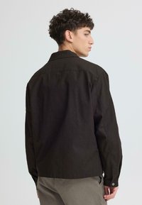 Chemise noire à manches longues avec col, poignets boutonnés et surpiqûres visibles. Le tissu semble lisse avec une coupe légèrement structurée.