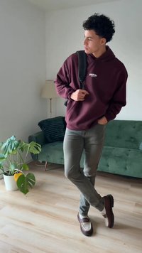 Vijoličast pulover s kapuco z logotipom, sive kavbojke, rjavi mokasini z belimi poudarki. Svetel lesen pod, zelena kavč in lončnica v ozadju.