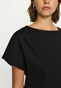 Fekete, rövid ujjú felső, széles nyakkivágással, puha anyagból készült. Látható varrásokkal és laza fazonnal rendelkezik, kiemelve a sima textúrát.