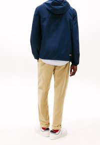 Námořnická bunda s kapucí a elastickými manžetami, v kombinaci s béžovými chino kalhotami a bílými teniskami. Červené ponožky viditelné nad okrajem bot.