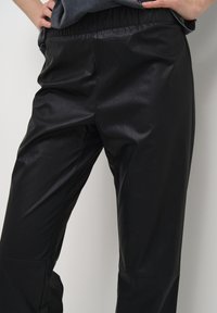 Černé kalhoty z umělé kůže s hladkým povrchem, elastickým pasem a zúženým střihem. Viditelné švy a minimalistický styl.