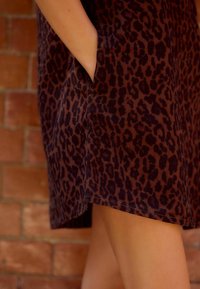 Braun leopardmuster Kleid mit einer weichen Textur, ausgestattet mit Seitentaschen und einem geraden Saum, vor einem Backstein-Hintergrund.