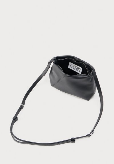 Čierna kožená crossbody taška s nastaviteľným popruhom, horným zipsom a viditeľnou bielou nálepkou vo vnútri na jednoduchom svetlom pozadí.