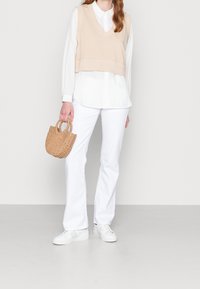 Suéter corto beige sobre una camisa blanca con cuello, combinado con pantalones blancos de campana y zapatillas blancas. Los accesorios incluyen un pequeño bolso de mano tejido.