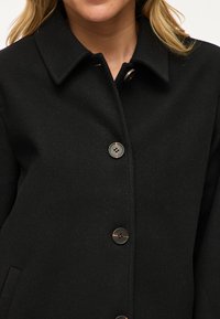 Žena nosi čierny kabát s golierom a štyrmi viditeľnými gombíkmi vpredu, ukazujúc hornú časť trupu a časť blond vlasov.