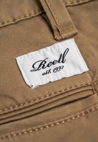 Brauner Stoff mit einem weißen Etikett, das aufgenäht ist und den Text "Reell est. 1997" in Kursivschrift zeigt, mit sichtbaren Stichdetails um das Etikett.