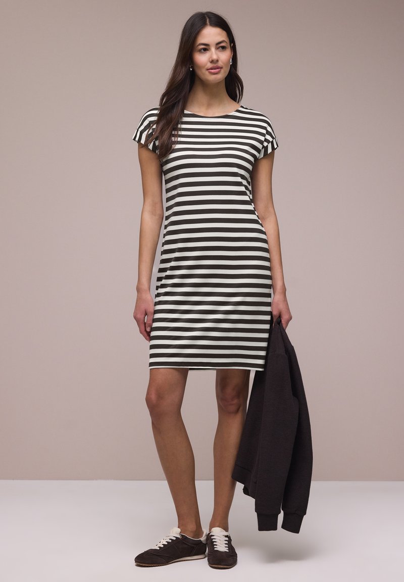 Femme portant une robe mi-longue à rayures horizontales noires et blanches, tenant une veste noire et portant des baskets noires avec des lacets blancs.