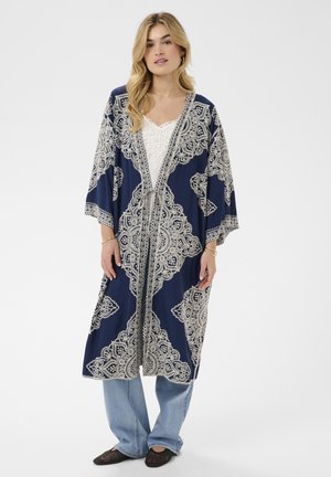 Woman wearing a white lace top, blue patterned long kimono-style cardigan, light blue jeans, and black woven shoes, standing against a plain background.