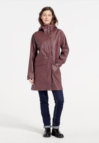 Burgunderfarbene wasserdichte Regenjacke mit Kapuze, Knopfverschlüssen und Seitentaschen, getragen über dunklen Jeans und Stiefeletten. Glatte, glänzende Textur.