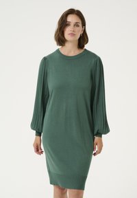 Robe en tricot verte avec de longues manches ballon plissées, un col rond et une jupe légèrement fuselée. Texture lisse avec des accents côtelés.