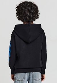 Felpa nera con cappuccio e grafica blu sulla manica sinistra, realizzata in tessuto morbido, abbinata a jeans in denim azzurro chiaro.