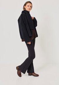 Giacca nera corta in tessuto morbido, indossata sopra un maglione marrone. Pantaloni neri aderenti e stivaletti marroni. Design semplice, hardware minimale.