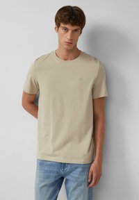 Beige katoenen t-shirt met korte mouwen, ronde hals en een klein logo op de borst, gecombineerd met lichtblauwe jeans.