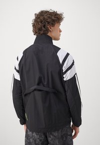 Veste légère noire avec des accents rayés blancs et noirs, col haut et poche au dos. Tissu texturé avec une coupe décontractée.