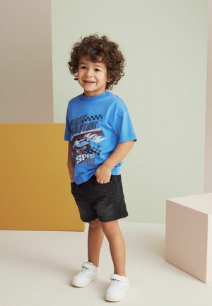Blaues Grafik-T-Shirt mit Rennsport-Thema, schwarze Shorts und weiße Sneakers. Die lockigen Haare des Kindes und das Lächeln sind auffällige Merkmale.
