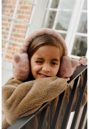 Měkké růžové fleecové uši s medvědími akcenty, které nosí dítě v béžovém texturovaném kabátu, opírající se o černý zábradlí, uvnitř světlé místnosti.