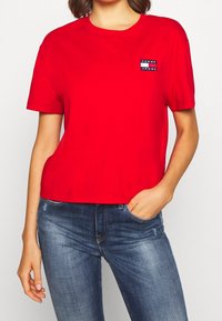 Kobieta w jasnoczerwonej koszulce Tommy Jeans z małą naszywką logo na piersi, w połączeniu z niebieskimi dżinsami.