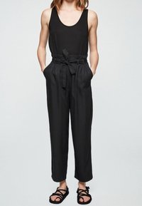 Combinaison noire avec des jambes larges et une ceinture nouée à la taille. Dotée d'un col arrondi et de poches. Assortie à des sandales plates noires.