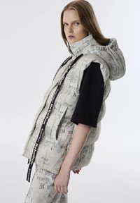 Dikke mouwloze vest in lichtbeige met een capuchon, voorzien van zwarte graffiti tekst en branding, en verstelbare trekkoorden bij de hals.