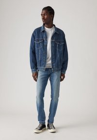 Jachetă din denim de culoare albastru închis cu două buzunare pe piept, asortată cu blugi albastre deschise și teniși negri cu tălpi și sireturi albe.