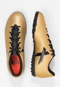 Kultaiset synteettiset jalkapallokengät mustilla yksityiskohdilla, teksturoitu päällinen, mustat nauhat ja punainen pohjallinen, joissa on erottuva logo ja kevyt rakenne.