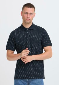 Polo noir avec un motif texturé comprenant de petites formes géométriques bleues, patte à deux boutons et col contrastant.