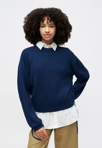 Sweater tricot azul-marinho com riscas, com decote redondo e corte relaxado, sobre uma camisa branca com riscas. Apresenta laços laterais e mangas longas.