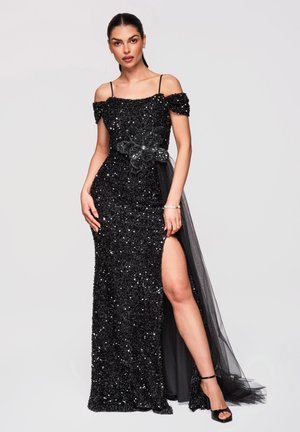 Femme portant une robe de soirée noire à sequins, épaules dénudées, fente haute et ornement floral, associée à des talons noirs à bride à la cheville.