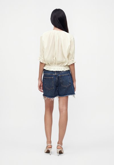 Femme aux longs cheveux foncés portant un chemisier crème, un short en jean bleu foncé effiloché et des sandales beiges à talons hauts, debout dos à la caméra.