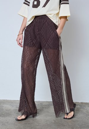 Donna che indossa pantaloni larghi di pizzo nero trasparente con strisce bianche laterali, tacchi neri aperti sulle dita dei piedi e una maglietta sportiva color crema con numero e strisce.