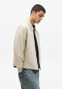 Beige jas met een puntige kraag, ritssluiting, geribde manchetten en zijzakken, gecombineerd met blauwe denim jeans. Lichaam zijwaarts gericht.