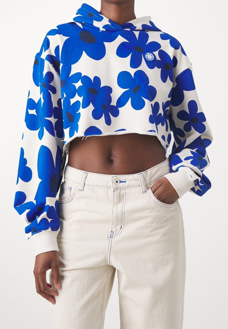 Sudadera recortada en blanco con un patrón floral azul, hecha de tela suave. Presenta una capucha con cordones y puños acanalados. Combinada con jeans de color claro.