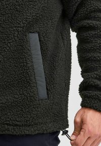 Fleece jacket in dark green with a textured surface, a black panel pocket, and adjustable elastic hem with toggle.