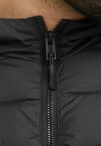 Black padded jacket featuring a zip closure, textured fabric with a grid pattern, and a stand-up collar. Close-up view focuses on the zipper.