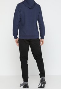 Uomo in piedi di spalle, che indossa un hoodie blu navy, pantaloni jogger neri e sneakers grigie Puma, su uno sfondo bianco semplice.